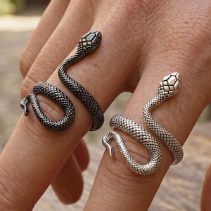 May include: Two wrap-around snake rings worn on fingers. One ring is finished in a dark, oxidized gunmetal grey, while the other is a bright, polished silver tone. Both rings feature detailed scale textures and a coiled design that wraps around the finger.