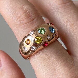 May include: A wide, polished gold-tone band ring featuring a scattered arrangement of multi-colored, round-cut gemstones. The stones include clear, yellow, green, blue, orange, and red crystals embedded directly into the smooth, rounded metal surface.