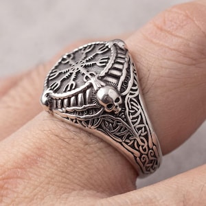 May include: A silver-toned metal ring featuring a circular face engraved with a Vegvisir symbol. The band displays intricate knotwork patterns on the sides and includes a small, detailed skull motif positioned directly below the central emblem.