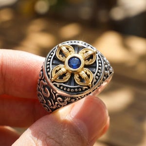 May include: A silver ring featuring a circular face with a gold-toned vajra symbol centered around a small, round, deep blue gemstone. The band and the outer edge of the ring face are decorated with intricate, raised, dark-oxidized scrollwork patterns.