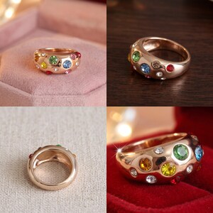 May include: A rose gold-toned ring featuring a wide, polished band embedded with multiple small, round, multi-colored gemstones including green, yellow, blue, red, and clear stones scattered across the surface of the metal band.