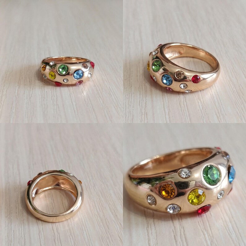 May include: A wide gold-tone metal ring featuring a polished finish and an array of embedded round multi-colored gemstones. The stones include clear, green, blue, yellow, and red hues scattered across the surface of the thick, rounded band.