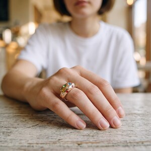 May include: A wide gold-toned ring featuring a domed surface embedded with multiple small, round, multi-colored gemstones including blue, red, yellow, and green stones arranged across the polished metallic band.
