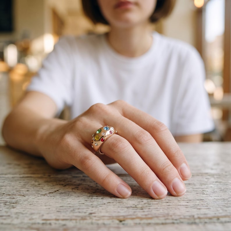 May include: A wide gold-toned ring featuring a domed surface embedded with multiple small, round, multi-colored gemstones including blue, red, yellow, and green stones arranged across the polished metallic band.