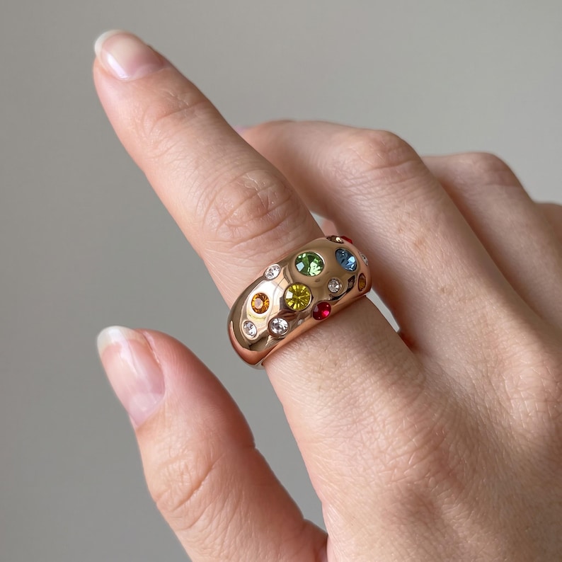 May include: A wide, polished gold-tone band ring featuring a scattered arrangement of multi-colored, round-cut gemstones. The stones include clear, yellow, green, blue, orange, and red crystals embedded directly into the smooth, rounded metal surface.