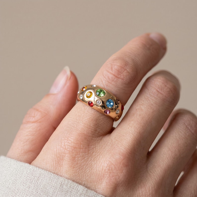 May include: A wide gold-toned band ring featuring a smooth, rounded surface embedded with multiple small, round, multi-colored gemstones including green, blue, red, orange, and clear crystals arranged in a scattered, asymmetrical pattern.