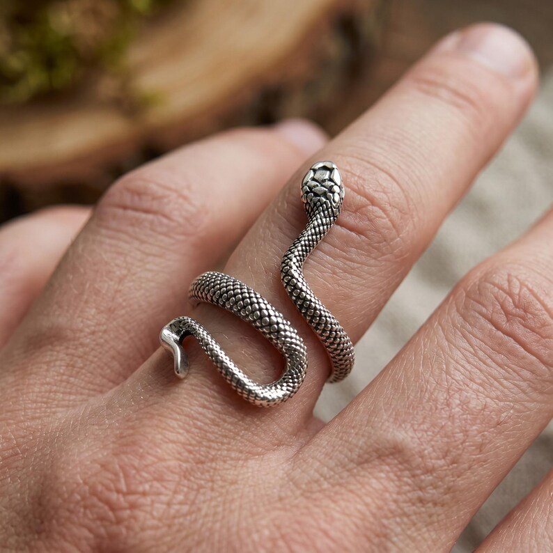 Pode incluir: Um anel de metal prateado em forma de serpente enrolada com texturas de escamas detalhadas. A cabe&ccedil;a da serpente repousa sobre o dedo superior, enquanto o seu corpo envolve o dedo num design em espiral, criando um aspeto de anel aberto.