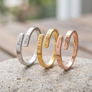 Puede incluir: Tres anillos abiertos en tonos plata, oro y oro rosa. Cada anillo presenta dos extremos planos y cuadrados grabados con texto. El anillo plateado dice Abel y Mark J, el dorado dice BE RAW y STAY SALTY, y el de oro rosa dice Wells e Indy.