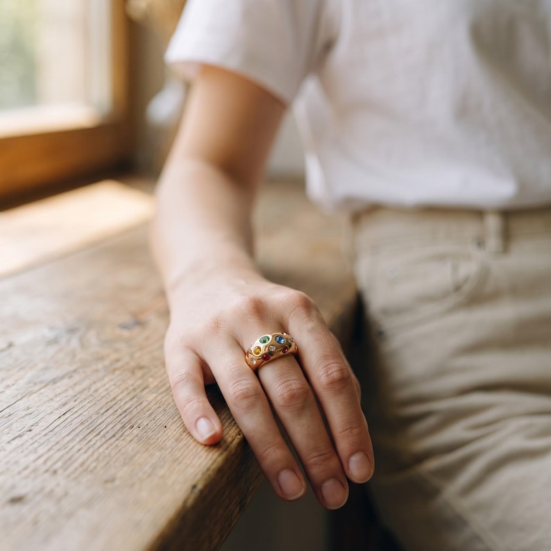 May include: A wide gold band ring featuring a textured, organic surface with multiple flush-set, multi-colored gemstones including blue, green, yellow, and red stones embedded across the rounded metal surface of the jewelry piece.