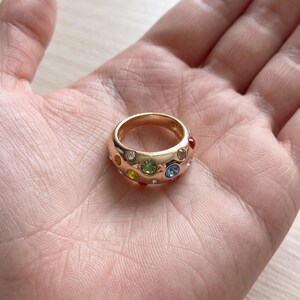 May include: A gold-toned dome ring featuring a variety of embedded round, multi-colored gemstones including green, blue, yellow, red, and clear stones set across the polished surface of the wide metal band.