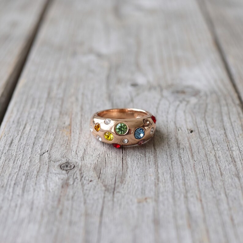 May include: A rose gold-toned band ring featuring a wide, rounded profile embedded with multiple small, circular faceted crystals in various colors including light green, blue, yellow, orange, red, and clear white, arranged in a scattered pattern.