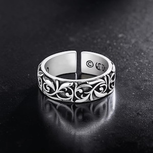 Puede incluir: Anillo plateado con diseño abierto. El anillo presenta un intrincado patrón floral repetitivo alrededor de la banda. El interior del anillo es liso y pulido. El anillo se muestra sobre una superficie oscura.