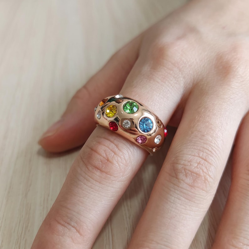 May include: A wide, polished rose gold-tone band ring featuring a scattered arrangement of round, multi-colored faceted crystals. The embedded stones include vibrant shades of green, blue, yellow, red, pink, and clear white, set flush into the metal surface.