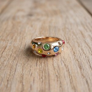 May include: A gold-toned metal ring featuring a wide, rounded band embedded with multiple small, circular, multi-colored gemstones. The stones include shades of green, blue, yellow, orange, red, pink, and clear white, set into the polished surface.