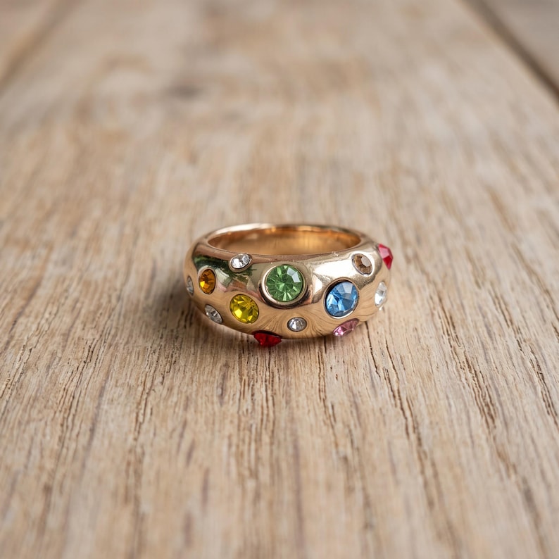 May include: A gold-toned metal ring featuring a wide, rounded band embedded with multiple small, circular, multi-colored gemstones. The stones include shades of green, blue, yellow, orange, red, pink, and clear white, set into the polished surface.