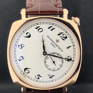 Puede incluir: Reloj de lujo con caja dorada y esfera crema. Cuenta con números arábigos negros, un pequeño dial de segundos y una correa de cuero marrón. Se puede ver la marca "VACHERON CONSTANTIN GENEVE".