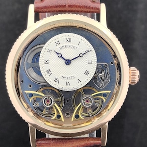 May include: A gold-toned wristwatch with a brown leather strap. The watch face features Roman numerals, the brand name "Breguet", and the number "1375". The watch has a visible mechanical movement and a blue background.