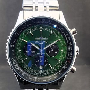 Könnte beinhalten: Eine Breitling Navitimer-Uhr aus Edelstahl mit grünem Zifferblatt. Die Uhr verfügt über mehrere Hilfszifferblätter, eine Datumsanzeige und eine Tachymeterskala. Die Uhr hat ein silbernes Armband und eine schwarze Lünette. Der Text "BREITLING 1884" ist sichtbar.