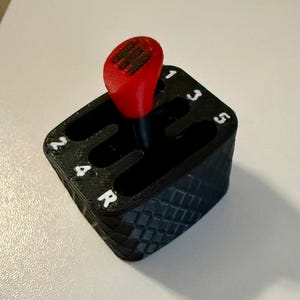 May include: A black 3D-printed gear shift knob with a red top. The knob has a diamond pattern on the sides and white numbers and letters indicating the gear positions. The gear shift is for a car.
