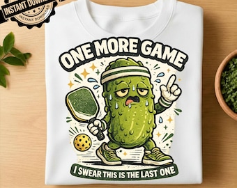 May include: A white t-shirt featuring a graphic of a green, sweating pickle character wearing a headband and sneakers. The pickle holds a pickleball paddle next to a yellow ball. Text reads: ONE MORE GAME I SWEAR THIS IS THE LAST ONE.