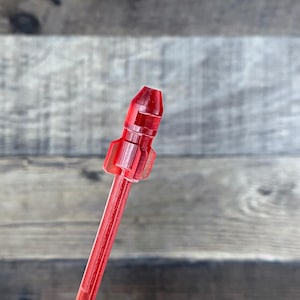 May include: A transparent red rocket-shaped cocktail stirrer. The stirrer has a long, thin stem and a rocket ship design at the top. The rocket has fins and a pointed nose. The stirrer is made of plastic and is angled diagonally.