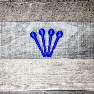 Op de afbeelding: Vier kobaltblauwe cocktailroerstaafjes met een ronde bovenkant en een lange, dunne steel. De roerstaafjes zijn in een waaier vorm gerangschikt tegen een achtergrond van een verweerd, grijs en wit houten oppervlak.