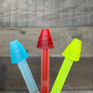 May include: Close-up of three colorful plastic dispensers. The dispensers are in shades of blue, red, and yellow-green. Each dispenser has a rectangular body and a conical top. The background is a blurred wooden surface.