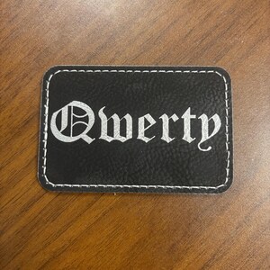 Puede incluir: Un parche rectangular negro con la palabra "Qwerty" en plata, con una fuente de estilo gótico. El parche tiene una superficie texturizada y costuras blancas alrededor de los bordes.