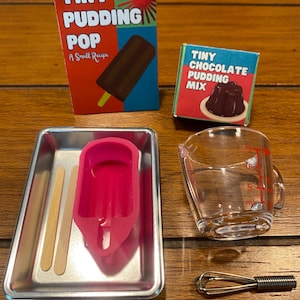 May include: A miniature set including a Tiny Pudding Pop box, a Tiny Chocolate Pudding Mix box, a rectangular metal tray with a pink silicone popsicle mold and two wooden sticks, a clear plastic measuring cup, and a small metal wire whisk.