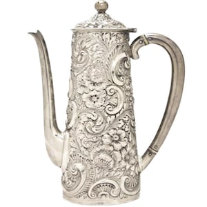 Peut inclure: Une cafetière en argent ornée de motifs floraux et de volutes complexes. Elle présente un long bec fin, une anse incurvée et un couvercle bombé avec un petit bouton. Les gravures détaillées couvrent toute la surface.