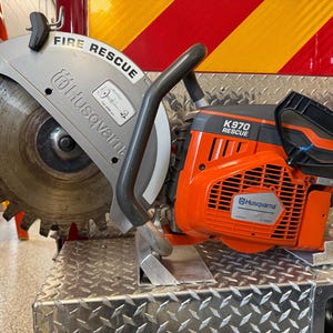 May include: A Husqvarna K970 Rescue power cutter featuring a large circular saw blade with prominent teeth, a grey metal blade guard labeled FIRE RESCUE, and a bright orange engine housing with black handles and control components.