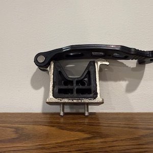 May include: A black and silver automotive part, likely a motor mount, featuring a black rubber component and a silver metal base with two bolts. A black lever is attached to the top.