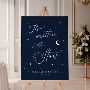 May include: A navy blue wedding sign on a wooden easel. The sign features white script that reads "It's written in the Stars" with the names Rebekah & Stuart and the date 12-12-27. The design includes stars and a crescent moon.