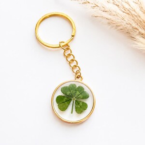 May include: A gold-toned metal keychain featuring a circular pendant with a clear resin center. Inside the resin is a single, pressed four-leaf clover with green leaves. The pendant is attached to a gold-toned split ring by a short metal chain.