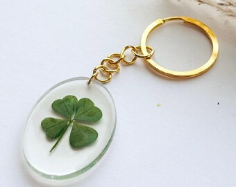 Può includere: Un portachiavi color oro con un ciondolo ovale. Il ciondolo racchiude un quadrifoglio in resina trasparente. Il portachiavi ha un anello e una catena dorati. Lo sfondo è bianco con alcune piante secche.