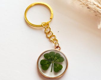 Può includere: Un portachiavi color oro con un ciondolo rotondo contenente un quadrifoglio. Il quadrifoglio è verde e racchiuso in resina trasparente. Il portachiavi ha un anello e maglie dorate.