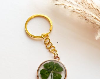 Può includere: Un portachiavi color oro con un quadrifoglio racchiuso in un ciondolo rotondo trasparente. Il portachiavi ha un anello e una catena dorati. Lo sfondo è bianco tenue con alcune piante secche.