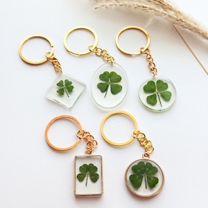 May include: A collection of six keychains featuring real four-leaf clovers encased in clear resin. The keychains have gold-colored metal rings and chains, with shapes including round, oval, and square. The clovers are a vibrant green against a white background.