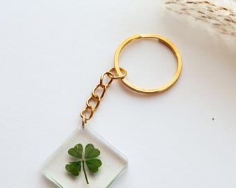 Può includere: Un portachiavi quadrato trasparente con un quadrifoglio racchiuso nella resina. Il portachiavi ha un anello e una catena color oro. Il quadrifoglio è verde e lo sfondo è bianco.