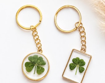 May include: Two gold-toned metal keychains featuring pressed four-leaf clovers encased in clear resin. One keychain has a circular frame, and the other has a rectangular frame. Both include a gold-toned split ring attached by a short chain.