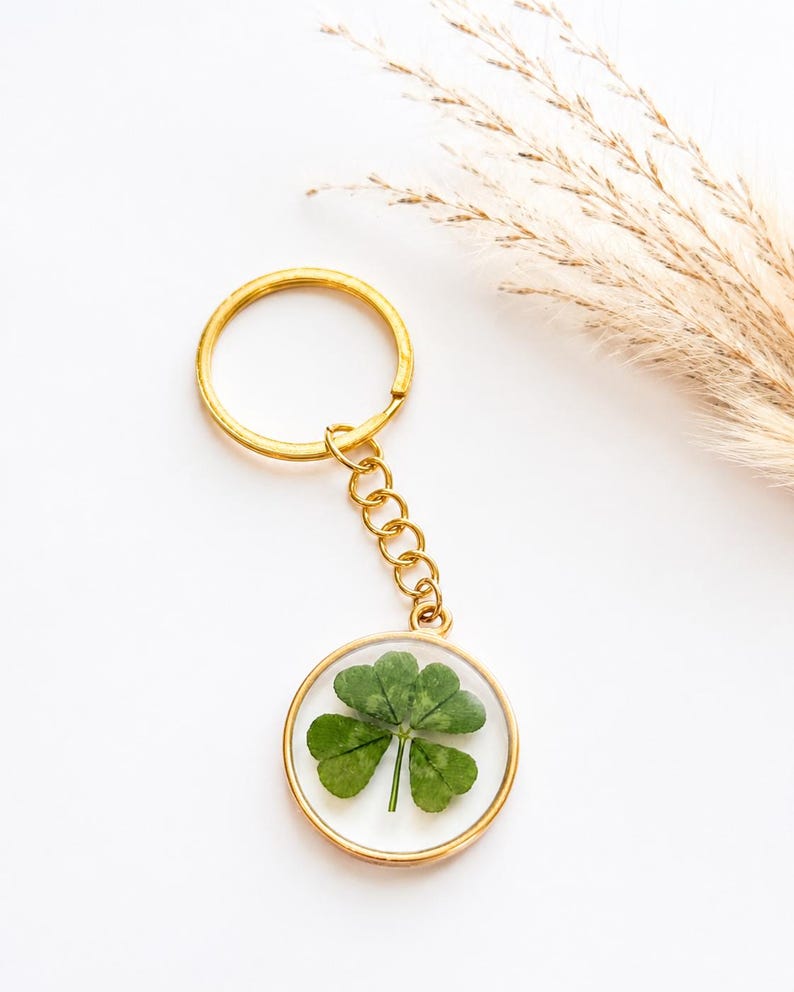 May include: A gold-toned metal keychain featuring a circular pendant with a clear resin center. Inside the resin is a single, pressed four-leaf clover with green leaves. The pendant is attached to a gold-toned split ring by a short metal chain.