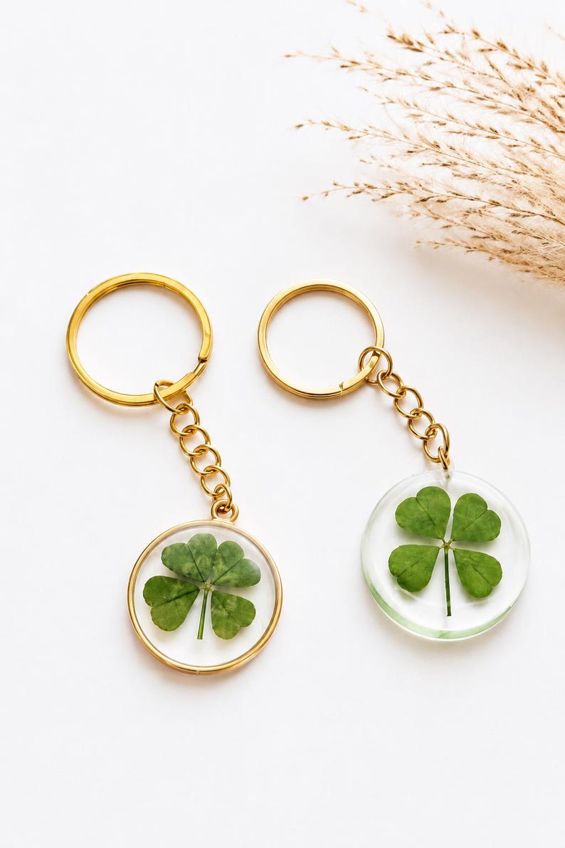 May include: Two circular keychains featuring four-leaf clovers encased in clear resin. One keychain has a gold-toned metal rim and chain, while the other is rimless with a gold-toned chain. Both include a circular metal split ring for keys.
