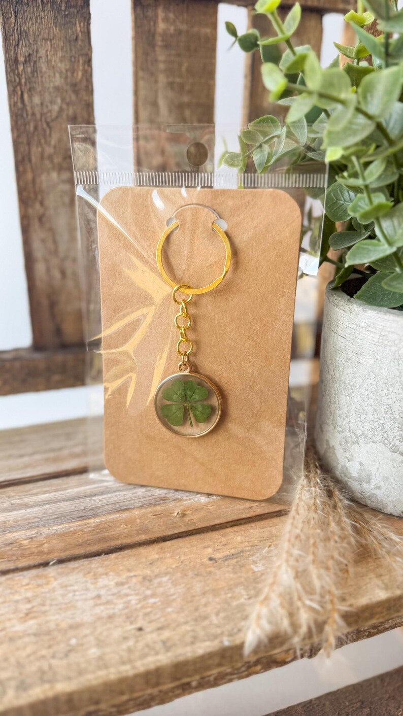May include: A gold-toned metal keychain featuring a circular pendant that encases a single pressed four-leaf clover. The keychain is attached to a rectangular brown card and enclosed in a clear plastic sleeve for protection and display.