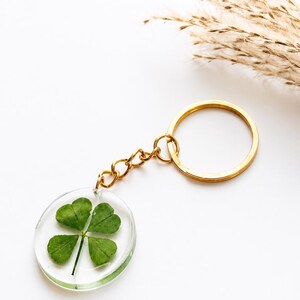 May include: A circular clear resin keychain containing a single pressed green four-leaf clover. The pendant is attached to a gold-toned metal jump ring and a matching gold-toned split key ring by a short gold-toned metal chain link connector.