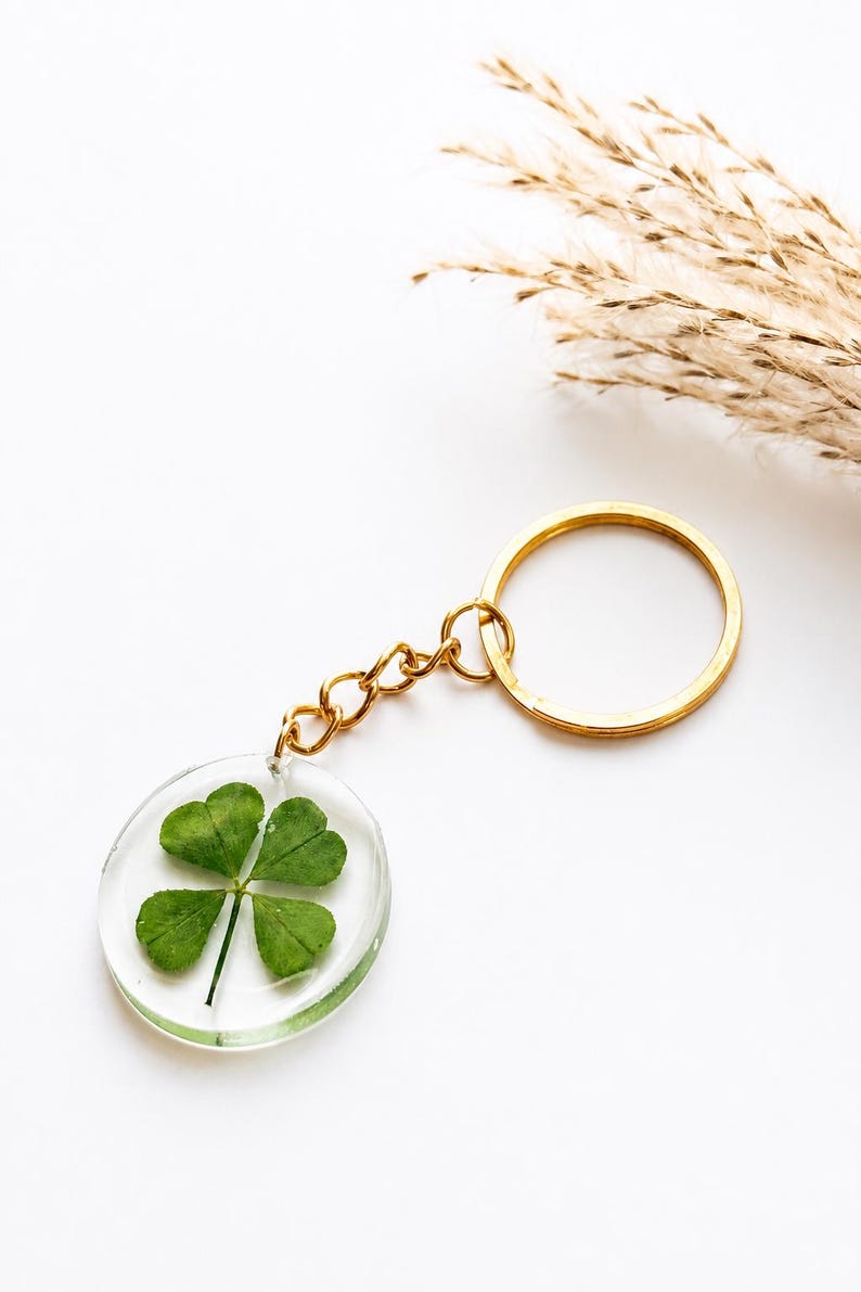 May include: A circular clear resin keychain containing a single pressed green four-leaf clover. The pendant is attached to a gold-toned metal jump ring and a matching gold-toned split key ring by a short gold-toned metal chain link connector.