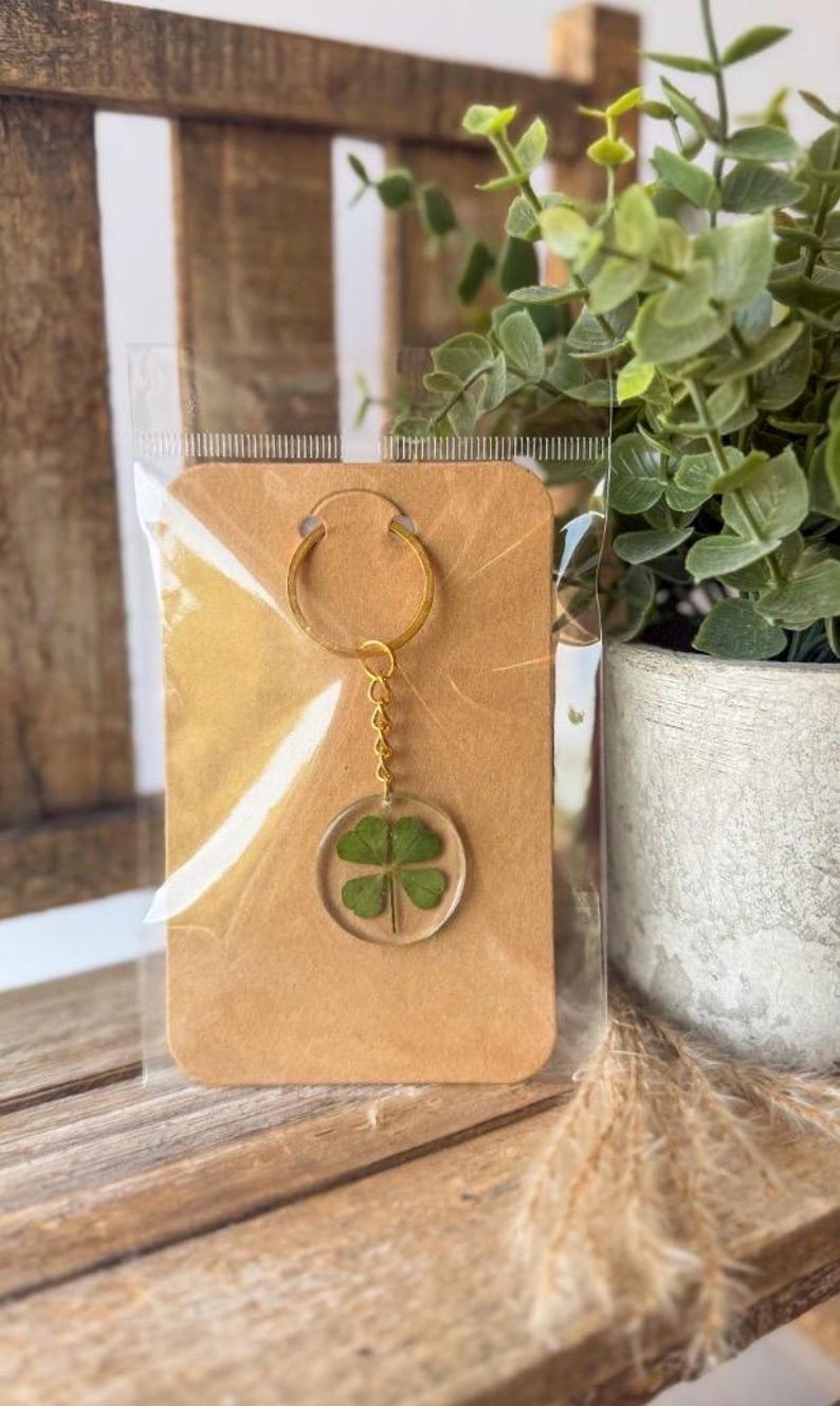 May include: A gold-toned metal keychain featuring a circular clear resin pendant that encases a real four-leaf clover. The keychain is attached to a rectangular brown cardstock display tag and is enclosed in a clear plastic protective sleeve.
