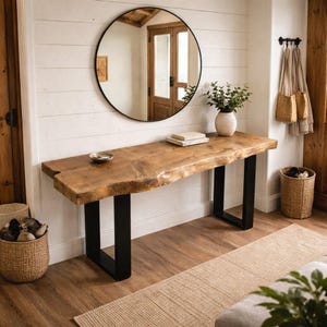 Puede incluir: Una mesa consola rectangular con una superficie de madera natural de borde irregular y dos patas de metal negro en forma de U. La mesa está colocada contra una pared blanca bajo un gran espejo redondo con un marco negro fino.
