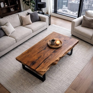Peut inclure: Table basse rectangulaire en bois avec bord naturel et finition marron foncé. La table repose sur des pieds en métal noir. Un petit plateau avec une tasse et une bougie est posé sur la table, sur un tapis gris clair.