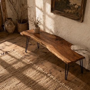 May include: A wooden bench with a live edge, featuring black hairpin legs. The bench is a warm brown colour and has a smooth surface. A neutral-toned throw is draped across the bench. The bench is approximately 150cm long.