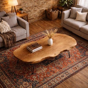 Op de afbeelding: Een houten salontafel met een organische vorm en haarspeldpoten. Op de tafel staan een vaas met droogbloemen en boeken. De tafel staat op een vloerkleed met een patroon in een woonkamer.